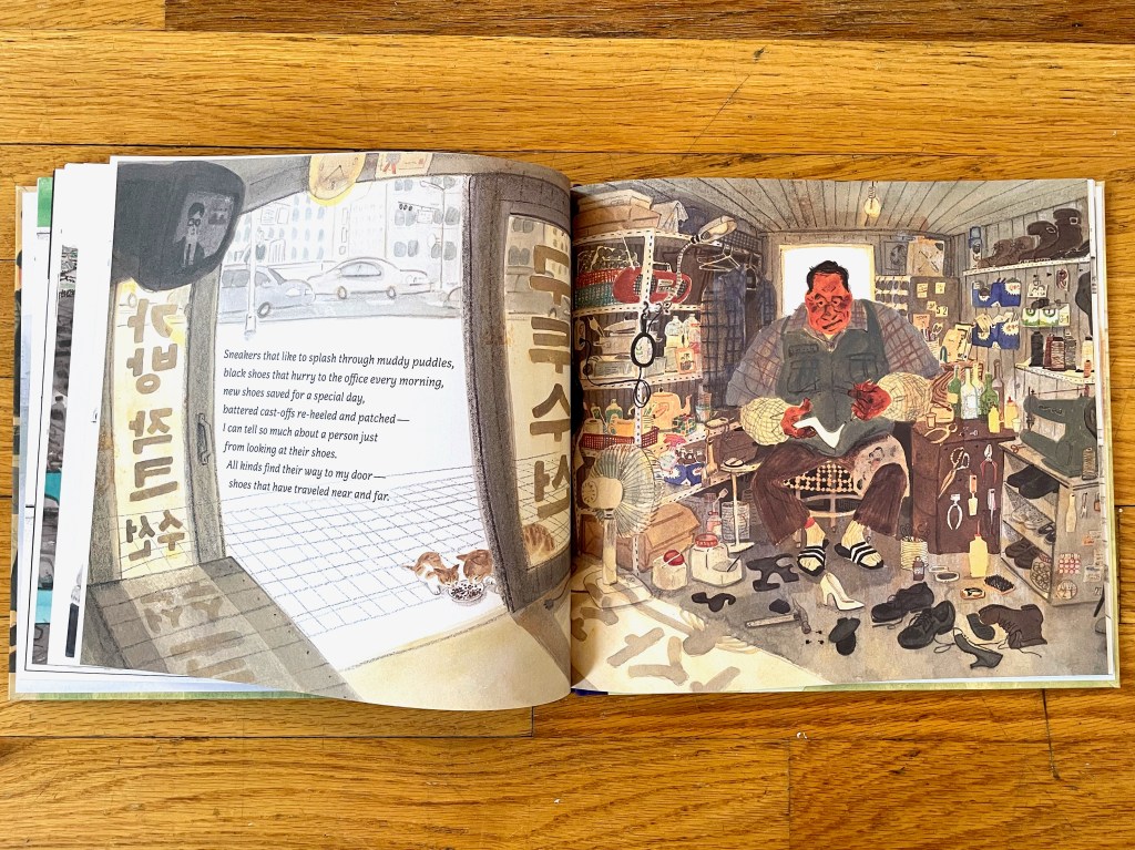The full spread shows inside the shop of a man working on repairing shoes. Two cats can be seen through the open door eating from a plate.