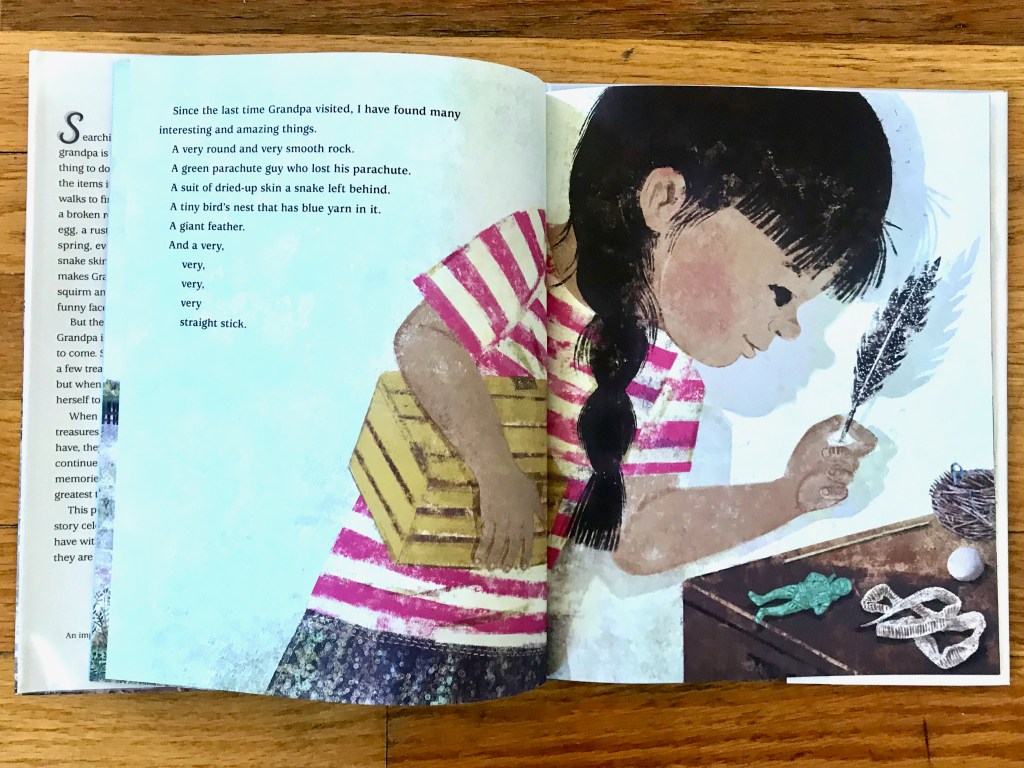 Little girl with a black braid and tan skin holds a yellow box in one arm while she places a black speckled feather onto a table holding a green figure, snake skin, egg, and part of a nest.
