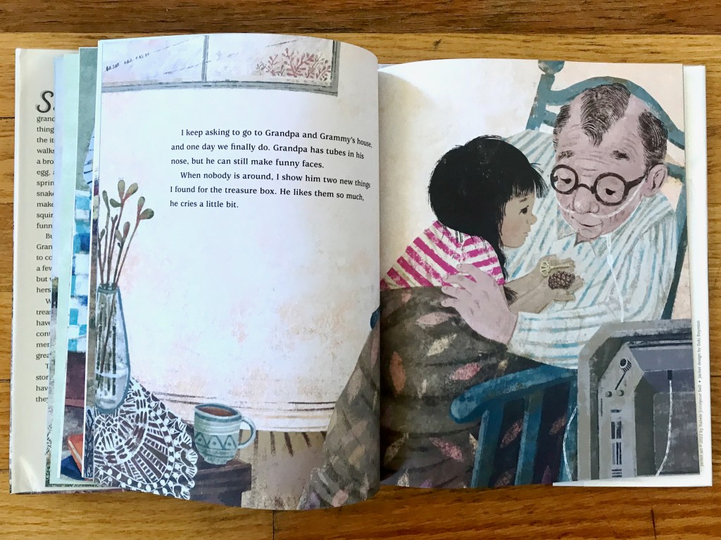 The grandpa sits in a rocking chair with a tube coming out of his nose. The little girl sits on his lap, wrapped in a blanket. She is holding a key and a pinecone for him to see.
