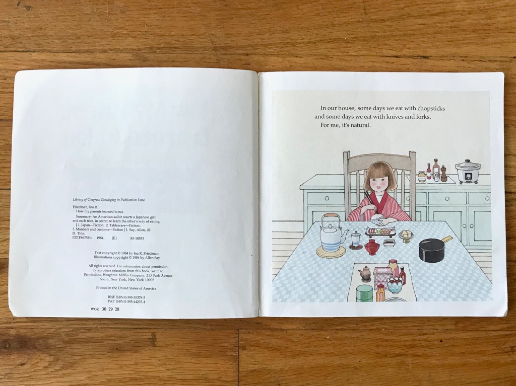 A little girl sits at a table in a kitchen wearing a red kimono, holding chopsticks and a bowl of rice.